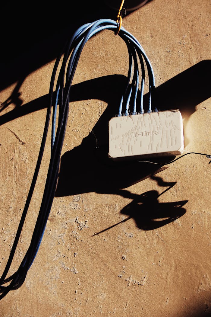 White electrical network adapter with various blue wires hanging on rough brown wall of building with bright sunlight and shadow